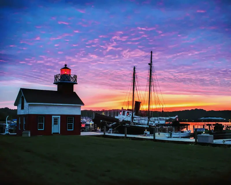 A vivid pink and purple sunset behind a red lighthouse and a schooner’s masts